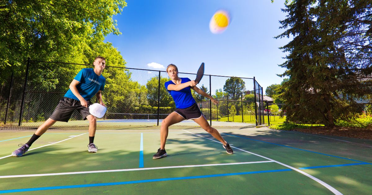Young male and female playing pickleball on a sunny day with racquets and ball in hand.