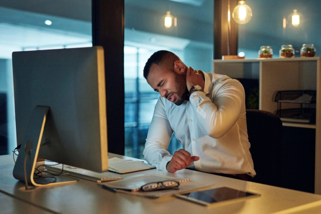 Photo of a man sitting at his desk holding his neck, demonstrating poor postural habits. Ergonomics can help prevent discomfort and injury from prolonged sitting.